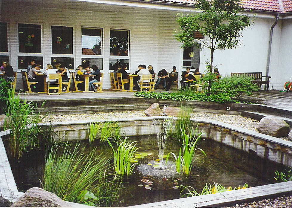 Innenhof mit Teich, Holzterrasse und lernenden Schülern auf Sitzgelegenheiten.